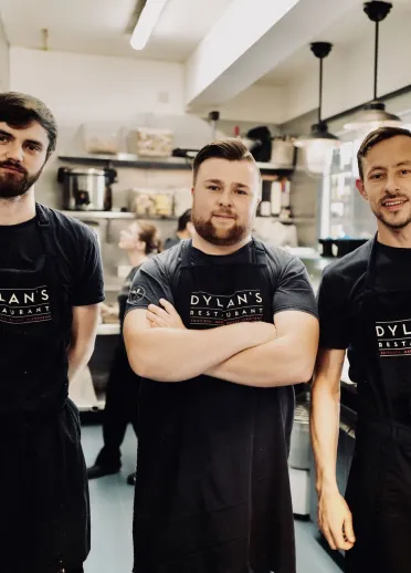 Tri aelod o staff yn gwisgo ffedogau du Dylan’s Restaurant yn sefyll mewn cegin fasnachol, gydag offer dur di-staen, silffoedd a goleuadau crog yn y cefndir.