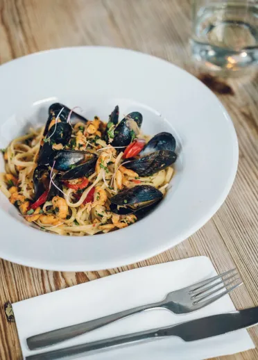 Pasta bwyd môr gyda chregyn gleision, corgimychiaid a phupur coch, wedi'i weini mewn powlen wen ar fwrdd pren, gyda chyllyll a ffyrc a gwydraid o ddŵr wrth ei ochr.