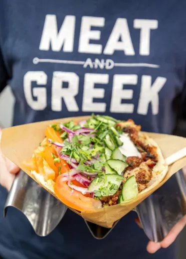 Person yn gwisgo “MEAT AND GREEK” T-shirt yn dal Greek-style flatbread Wedi'i lenwi â chig wedi'i grilio a salad.