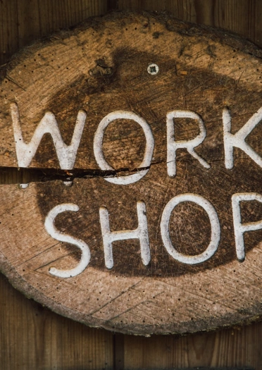 Y gair Workshop wedi’i gerfio i arwydd pren ar ddarn o risgl coeden.