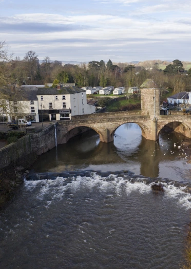 Pont gerrig hanesyddol yn croesi afon, gyda thai a siopau ar hyd glannau’r afon a chored yn y blaendir.