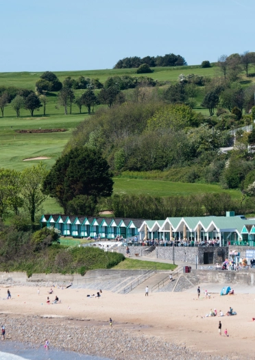 Golygfa o'r môr tuag at draeth tywodlyd gyda chytiau traeth gwyrdd.