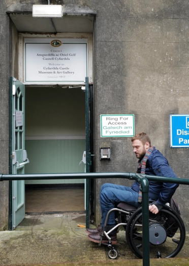Dyn mewn cadair olwyn yn defnyddio ramp i gael mynediad i adeilad. 