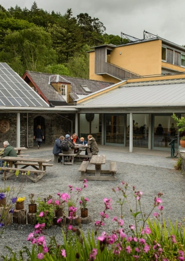 Pobl yn eistedd y tu allan i adeilad ar fyrddau picnic gyda blodau o’u hamgylch.