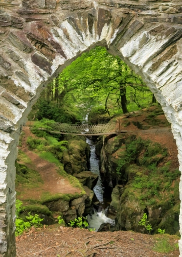 Rhaeadr coetir a phont gadwyn sy’n cael ei weld drwy fwa gothig.