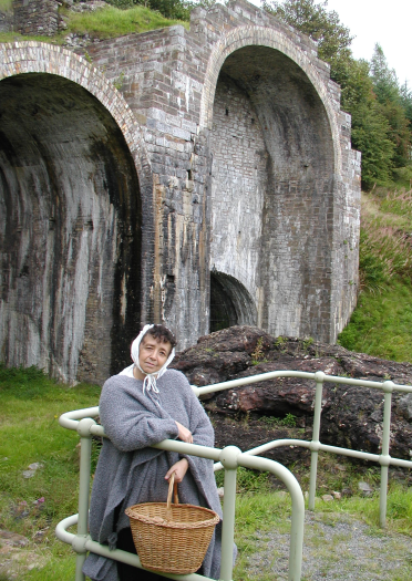 Gwraig mewn sgarff pen a chlogyn llwyd o flaen gweithfeydd diwydiannol.