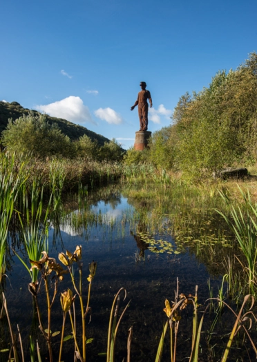 Cerflun metel o ddyn yn gwisgo het glöwr, ger llyn a choed gwyrdd.