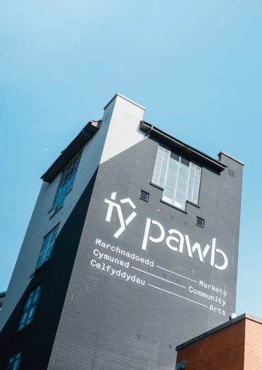 Llun o du allan canolfan gelfyddydol Tŷ Pawb – mae’r arwydd ar yr adeilad llwyd uchel yn dweud ‘marchnadoedd, cymuned, celfyddydau’. Mae'r awyr yn las ac mae baneri'n siglo yn y gwynt wrth ymyl yr adeilad.