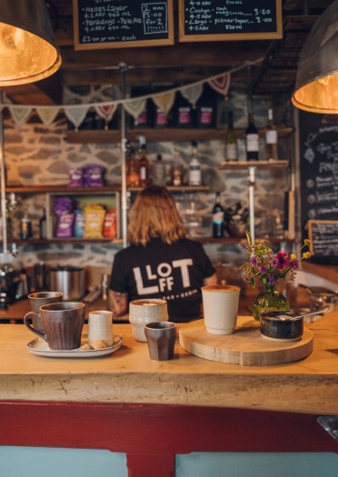 Aelod o staff tu ôl i far pren gyda choffi arno. Ar gefn crys-t y person mae enw'r bwyty - Llofft.
