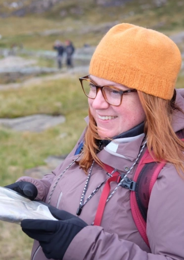 Person yn gwisgo sbectol, cot borffor a het oren yn dal map wrth fynydda.