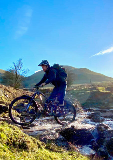 Dyn ar feic mynydd yn mynd drwy nant.