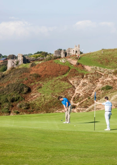 Golffiwr yn paratoi i bytio at y twll gyda golffiwr yn dal y faner yng Nghlwb Golff Pennard gyda Chastell Pennard yn y cefndir.