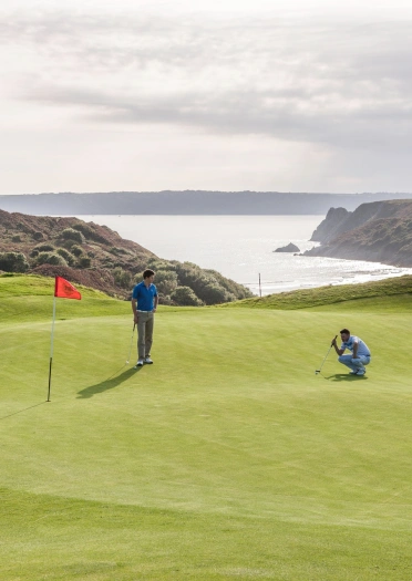 Dau golffiwr ar y grîn yng nghlwb golff Pennard gyda'r mynyddoedd a'r môr yn y cefndir
