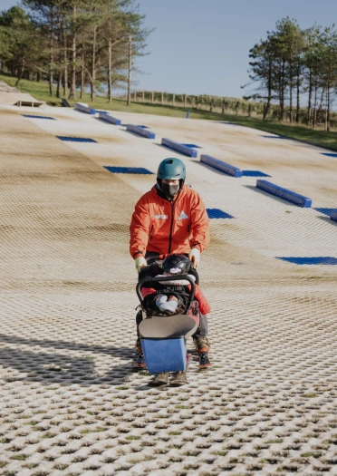 Plentyn mewn sled / pram sgïo wedi’i addasu yn cael ei wthio gan ddyn ar lethr sgïo sych.