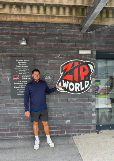 Dyn yn sefyll ger arwydd Zip World