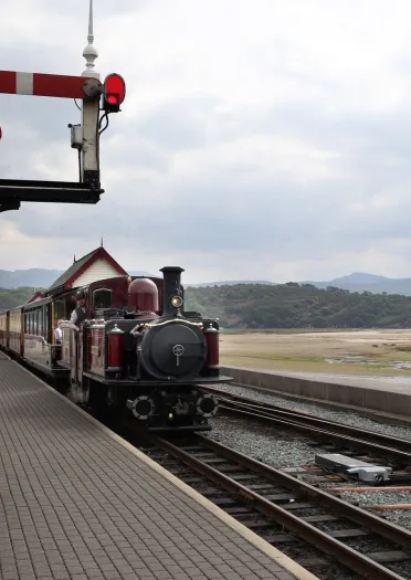 Locomotif stêm Double Fairlie a thrên mewn gorsaf reilffordd ger harbwr.