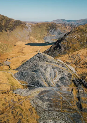 Yr olygfa o chwarel lechi ar ochr bryn gyda hen offer, adfeilion adeiladau, a thomenni llechi.