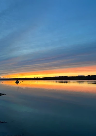 View from Y Felinheli across the Menai Strait showing still waters, a moored yacht and orange sunset reflections