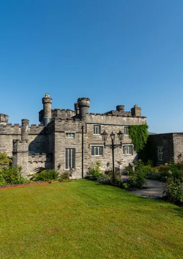 Castell carreg wedi’i hamgylchynu gan erddi a lawntiau wedi’u tirlunio, gyda borderi blodau lliwgar, lampau addurniadol a bryniau pell yn codi o dan awyr las glir.