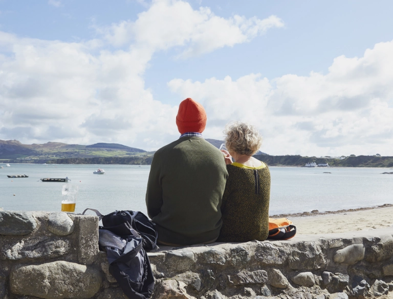 cefnau dau berson yn eistedd ar y wal, gyda golygfa o'r môr.