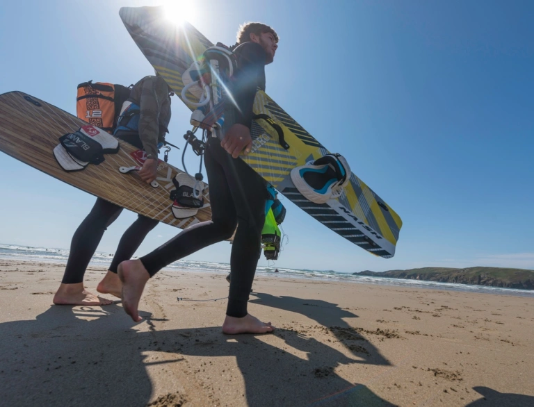 Dau berson yn cerdded ar draws traeth tywodlyd yn dal byrddau syrffio