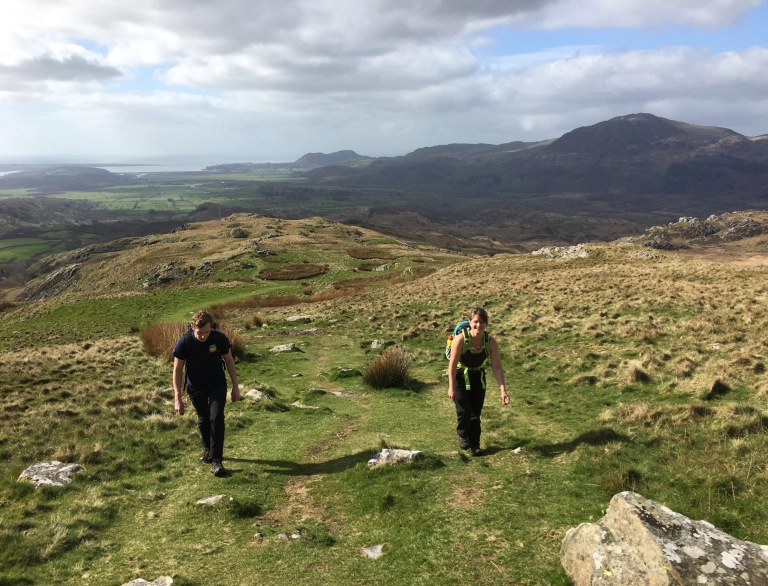 Dau berson yn cerdded ardal fynyddig gyda mynyddoedd ac arfordir yn gefndir.