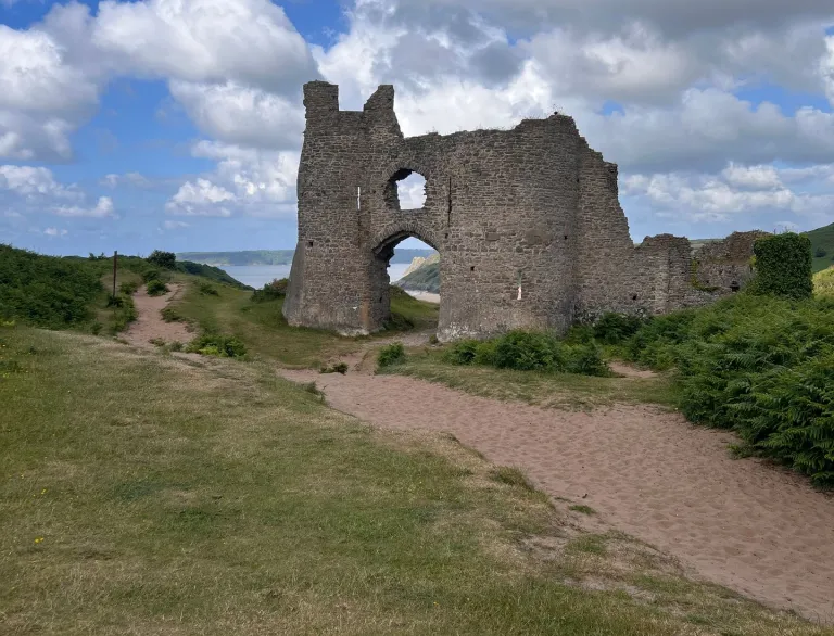 Adfeilion stondin castell hanesyddol yng nghanol glaswellt gwyrdd cyfoethog a llwybrau tywodlyd, o dan awyr las fywiog gyda chymylau fflyd.