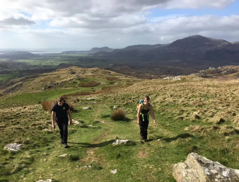 Dau berson yn cerdded ardal fynyddig gyda mynyddoedd ac arfordir yn gefndir.