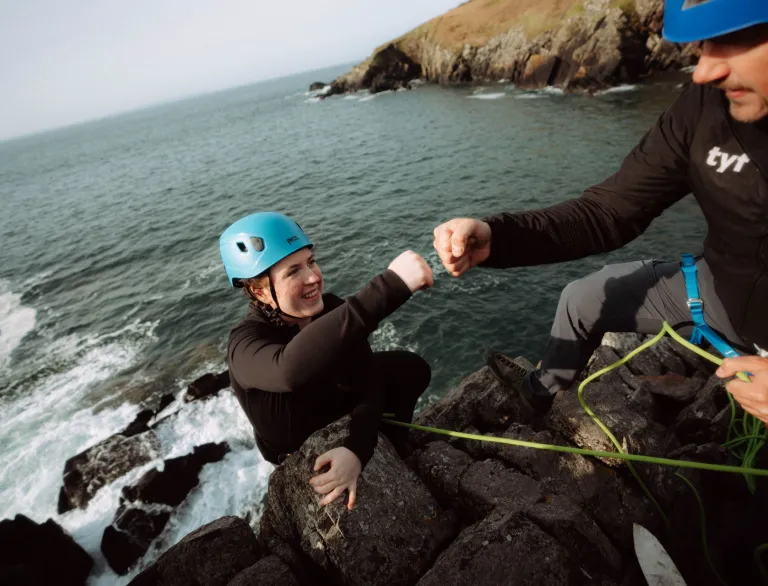 Dringwr a hyfforddwr, a’r ddau’n gwisgo helmedi, yn taro’u dyrnau yn erbyn ei gilydd ar wyneb craig gyda’r môr islaw.