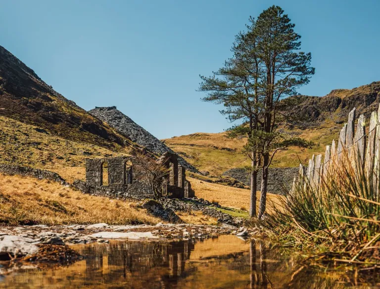 Hen chwarel lechi ar ochr bryn gydag adlewyrchiad adfeilion mewn dŵr llonydd. 