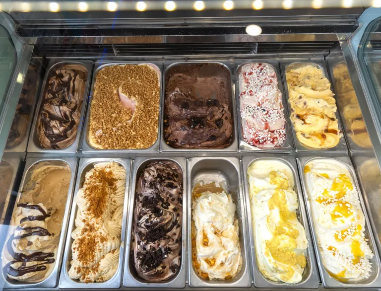 Detholiad o flasau gelato yn cael eu harddangos mewn hambyrddau metel y tu mewn i gownter oergell yn Zio's Gelateria.