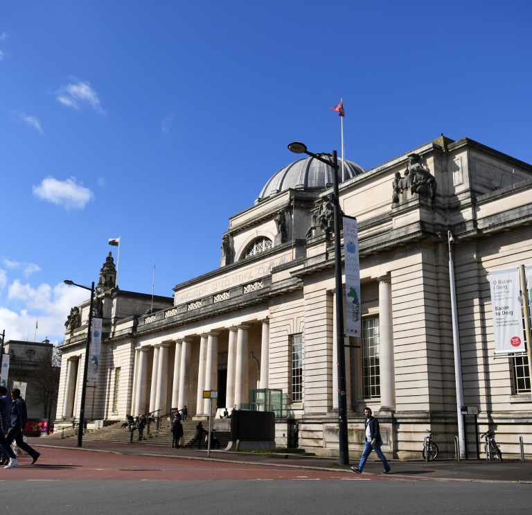 Tu allan i adeilad mawreddog gwyn Amgueddfa Genedlaethol Caerdydd ac awyr las yn gefndir.
