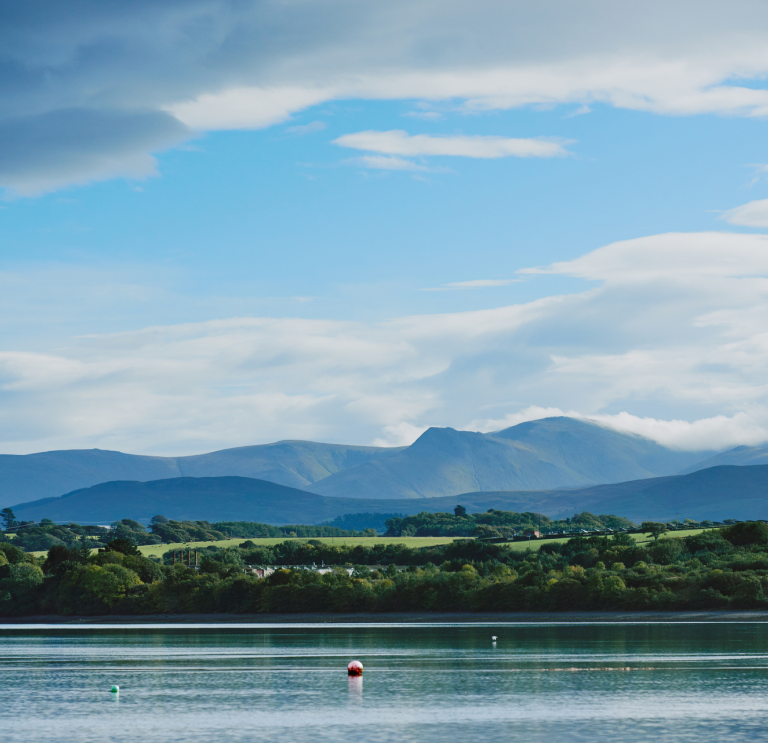 Llun o'r môr gyda choed a bryniau yn y pellter.