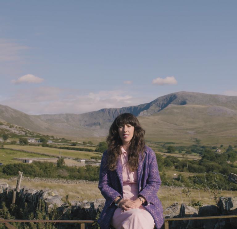 Landscape shot of singer Lisa Jen sat on a fence with the valley and surrounding mountains of Bethesda in the background.