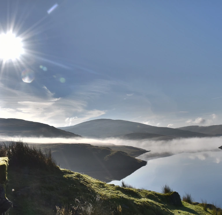 Rhywun yn edrych dros y mynyddoedd a llyn yn gynnar yn y bore.