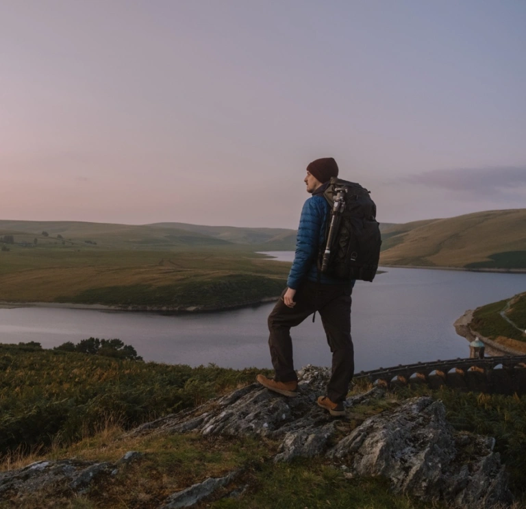 Dyn â bag ar ei gefn, yn sefyll ar ochr mynydd yn edrych allan ar draws tirwedd o fynyddoedd a llyn.