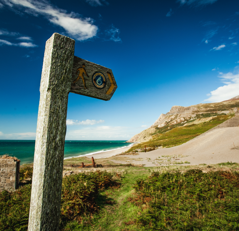 Arwydd llwybr arfordir Cymru gyda thwyni tywod a'r môr yn y cefndir