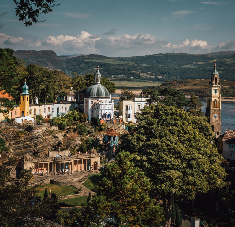 Golygfa o bentref Portmeirion o’r awyr gyda’r môr a’r mynyddoedd yn y cefndir.