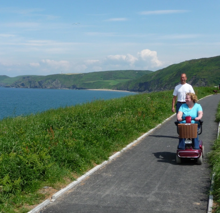 Menyw ar sgwter symudedd gyda dyn yn cerddedd ar ran darmac o lwybr arfordir cymru yn edrych allan i'r môr.