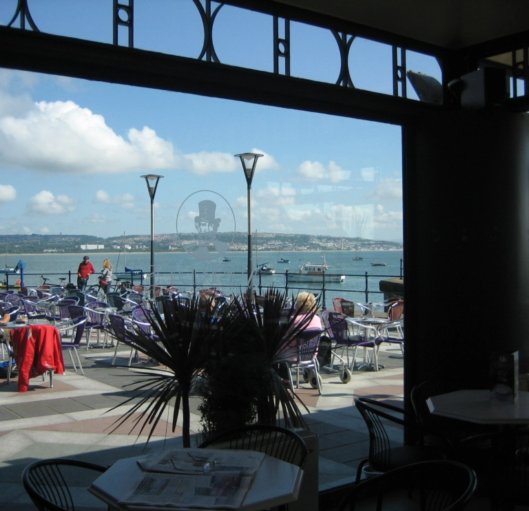 Lle eistedd awyr agored gyda’r môr y yn cefndir, a’r llun wedi’i dynnu drwy ffenest caffi.