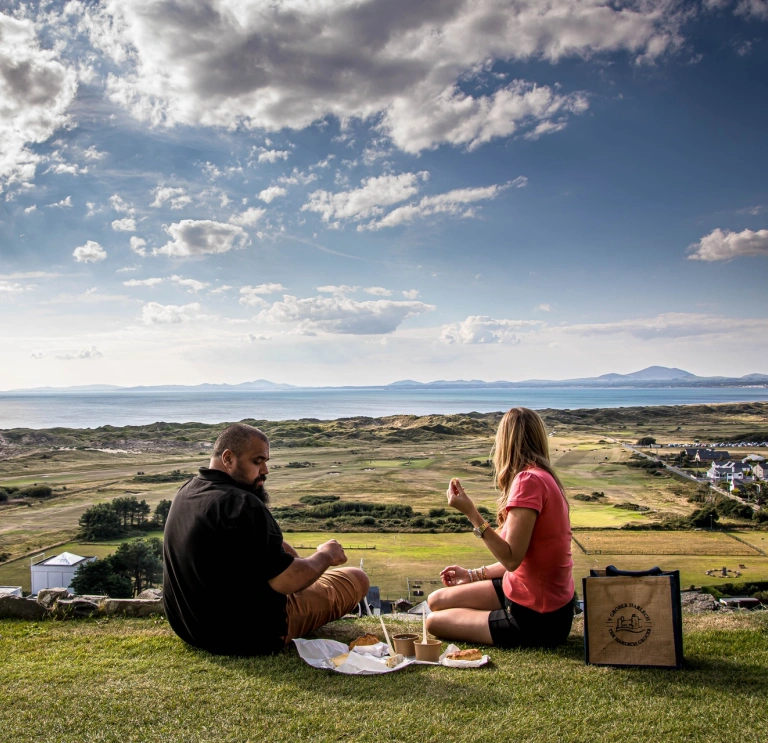 Dau berson yn cael picnic yn edrych dros cwrs golff.