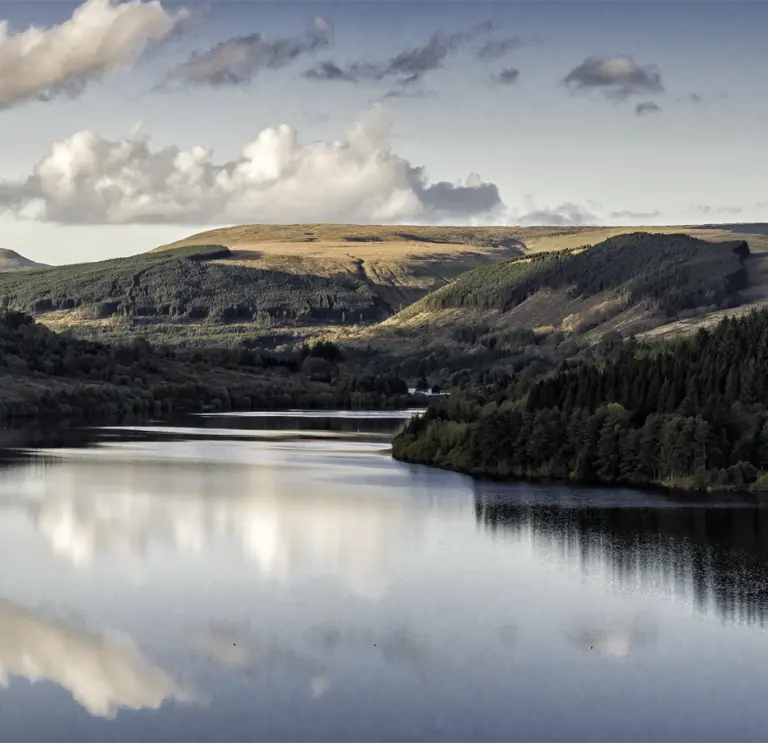 Adlewyrchiadau o awyr las gymylog ar wyneb llonydd y gronfa ddŵr, gyda choetir a mynyddoedd o’i chwmpas.