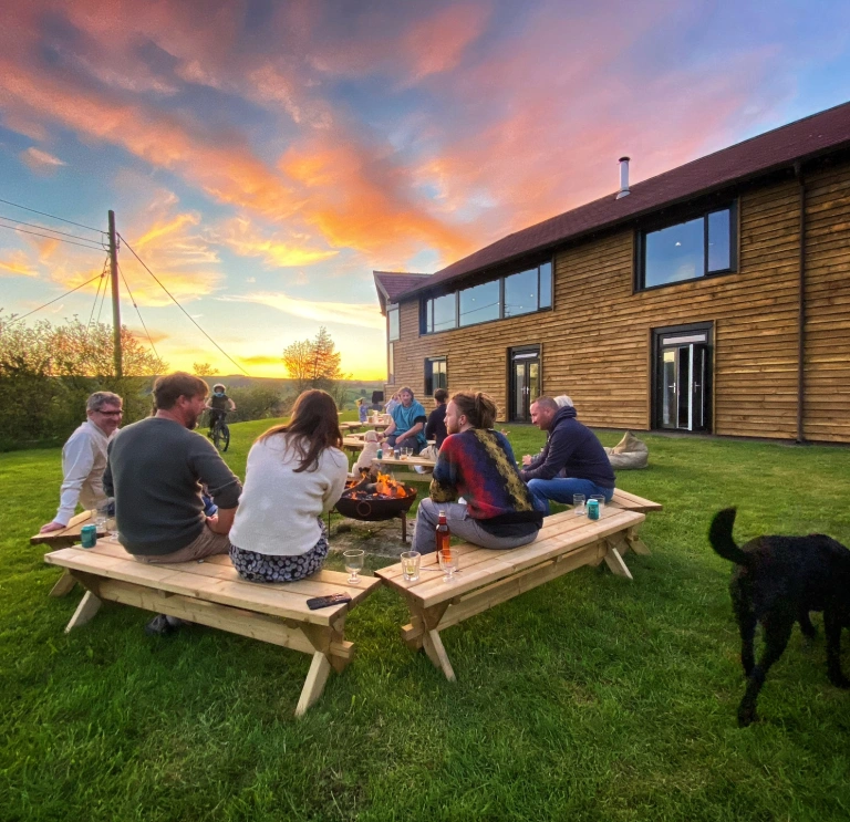 Grŵp o amgylch pwll tân y tu allan i adeilad arddull caban ar fachlud haul. 