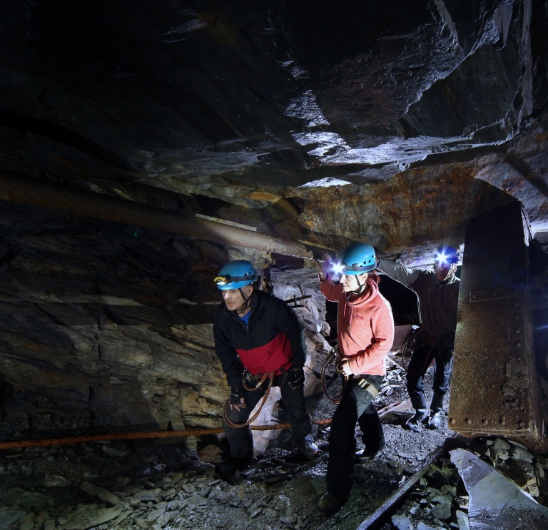 Grŵp o bobl yn gwisgo helmedi yn cerdded ar lwybr tanddaearol mewn chwarel lechi.