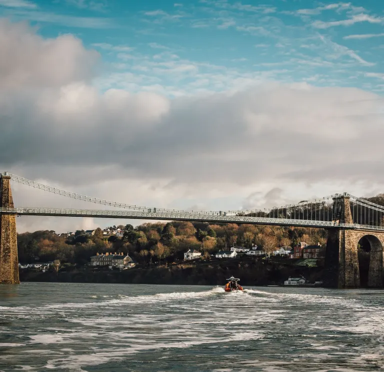 Llun o'r bont grog gyda choed a thai yn y cefndir a chwch ar Afon Menai