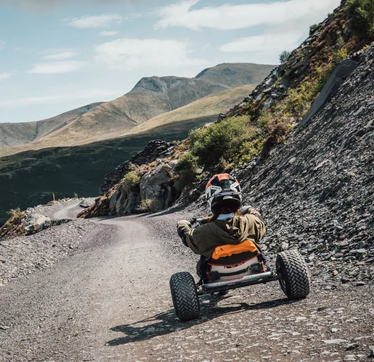 Person yn eistedd mewn go-kart yn teithio i lawr trac llechi llwyd gyda golygfeydd mynyddig yn y cefndir.