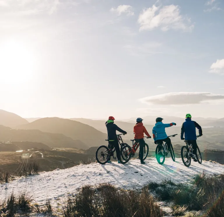 Roedd pedwar beiciwr yn dringo beiciau mynydd, yn sefyll yn edrych allan ar draws mynyddoedd eira.