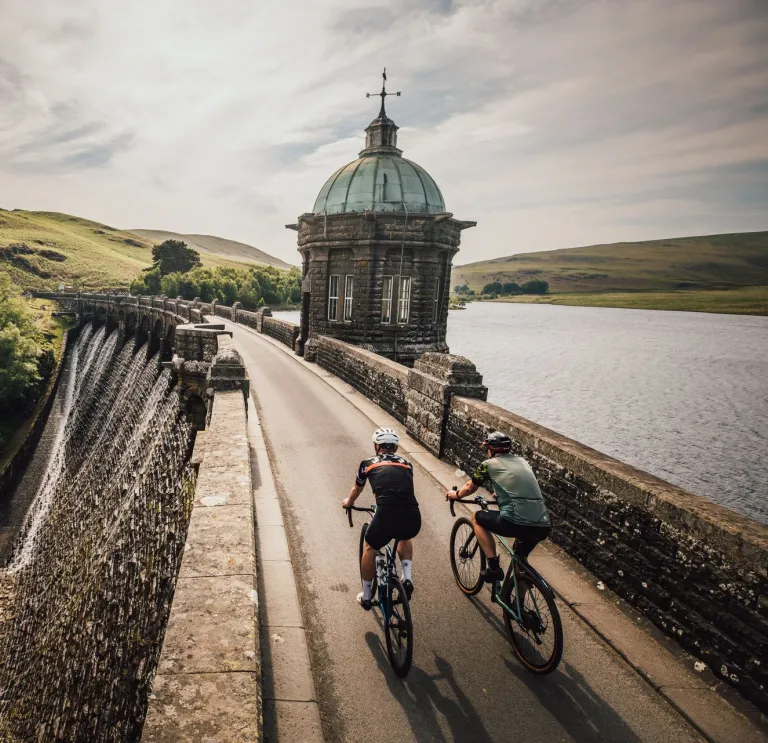 Dau feiciwr yn marchogaeth ochr yn ochr â chronfa ddŵr gyda chaeau gwyrdd i'w chwith a'r gronfa ddŵr i'r dde.