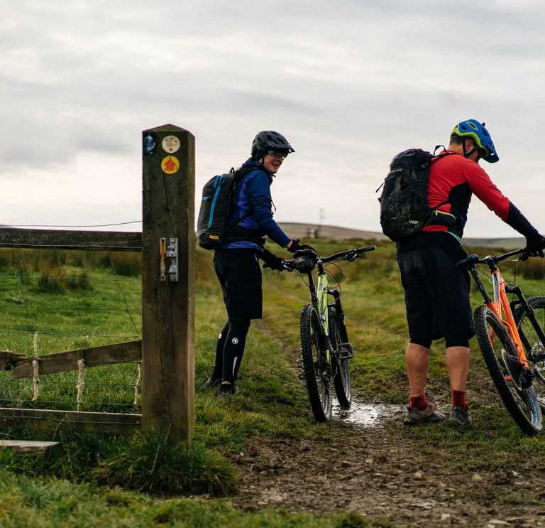 Dau berson wrth ymyl eu beiciau sy'n cael eu gosod ar y llawr, wrth giât i gae gydag arwydd yn nodi llwybr ceffyl ar y porth.