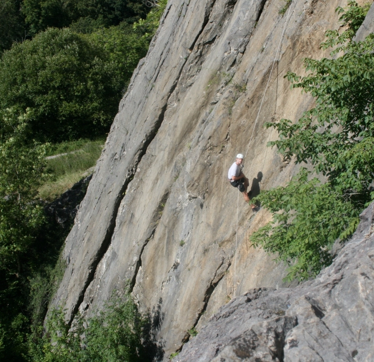 Dyn yn gwisgo offer diogelwch yn dringo wyneb craig.